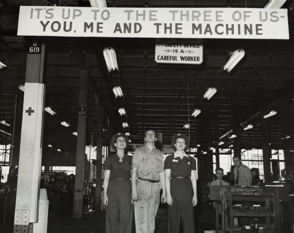 Factory workers posing under a sign at Internationl Harvester's St. Paul Works. Original caption reads: "The Production Drive Committee of Harvester's gun plant, composed equally of management and employee representatives, installed signs throughout the plant urging maximum cooperation by employees in the war effort. Here, left to right: Gladys Nimency, Vincent Huber, and Judith Benson are reading one of the signs." The signs above the workers read: "It's up to the three of us - You, Me, and the Machine;" and "The Best Safety Device is a Careful Worker."