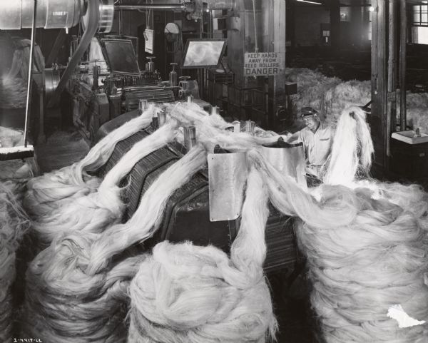 Factory worker feeding sisal fiber into a machine, possibly at International Harvester's McCormick Twine Mill. Original caption reads:  "The slivers fed into a second breakers receive another thorough combing to straighten fibers and make them smooth and even. Mirror in the background permits operator to make sure that the sliver is feeding properly into the machine."