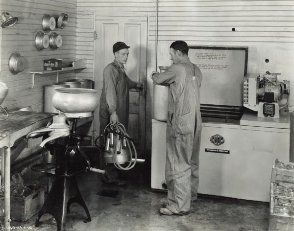 Two men are standing in a room with a cooler and milking equipment. Original caption identifies the equipment as "McCormick-Deering 6 Can Cooler and Mc[Cormick]-Deering Cream Separator and Milker."
