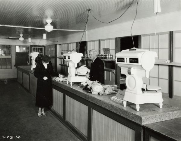 A man behind a counter weighs chickens on a countertop scale, and a women in a coat examines chickens on the counter. Some canned items are on a shelf behind the counter. Original caption reads: "McCormick-Deering Cooler Used for Storage of Dressed Poultry."