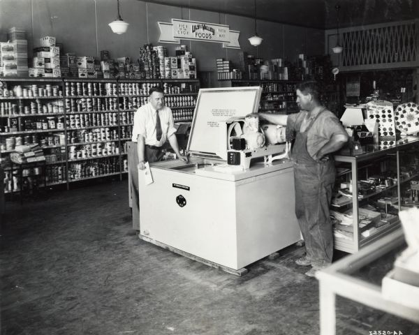 Two men, one in a tie, the other in overalls, stand next to a McCormick-Deering cooling unit on display in a grocery market. A sign on the back wall reads: "Lily of the Valley Foods; Pick of the Crop, Fresh Pakt." Original caption identifies the men as "(Left) P.W. Stevens, (Right) Melvin Butler."