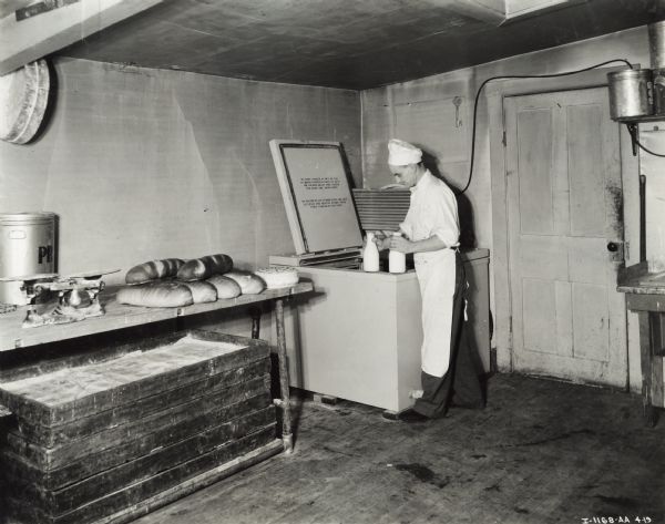 A man stands in front of a 4 can milk cooler with two milk jars in his hands at the Brauer Bakery.  Loaves of bread are on a table with a scale, a cake, and a container.