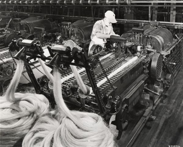 A man inspects a machine for spinning sisal, possibly at the McCormick Twine Mill. Original caption reads: "An operator is shown inspecting a spinning machine. This inspection is repeated constantly throughout the day to guard the quality of IH twine. Inset shows a full bobbin of twine being removed from the spinning machine for balling."