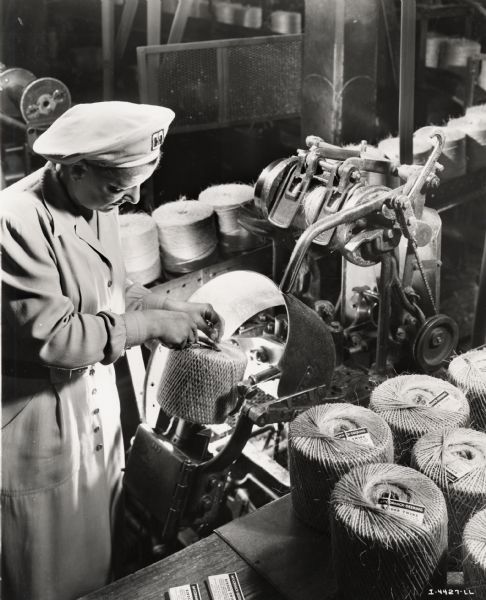 A female factory worker assembles a ball of twine, possibly at the McCormick Twine Mill. Original caption reads: "Tucking in the end of the twine after the criss-cross cover has been put on a ball of binder twine. The criss-cross cover allows the farmer to use the ball to the end, without its collapsing or tangling."