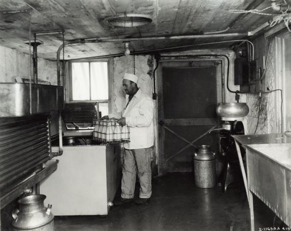 A man stands by a 6 can milk cooler holding a metal basket of milk jars at the Roland Schrap Dairy Farm. To the right of a door is a cream separator. A window is on the left.