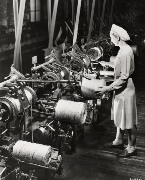 A female factory worker creates balls of twine, possibly at the McCormick Twine Mill. Original caption reads: "This scene shows a battery of balling machines making baler twine. Similar machines are used for balling binder twine. As a quality check, every ball of twine produced contains the machine operator's name."