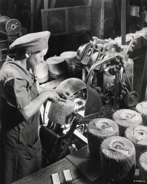 A female factory worker assembles a ball of twine, possibly at the McCormick Twine Mill. Original caption reads: "Tucking in the end of the twine after the criss-cross cover has been put on a ball of binder twine. The criss-cross cover allows the farmer to use the ball to the end, without its collapsing or tangling."