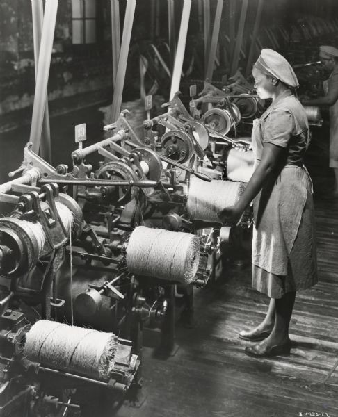 A female factory worker creates balls of twine, possibly at the McCormick Twine Mill. Original caption reads: "This scene shows a battery of balling machines making baler twine. Similar machines are used for balling binder twine. As a quality check, every ball of twine produced contains the machine operator's name."