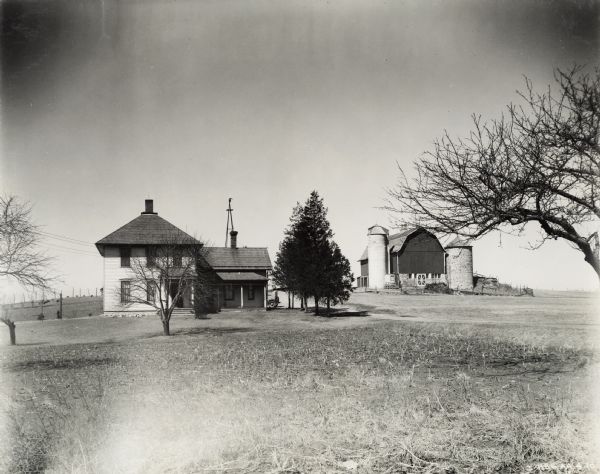 Exterior view of a two-story house with several trees on the farm of Fred Esser. On the right is a barn with two silos. On the far left in a fenced area on a small hill are chickens. A field of broken corn stalks is in the foreground. Mr. Esser owned a McCormick-Deering 6 can cooler.