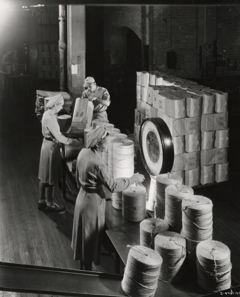 Female factory workers check the weight of bound twine, possibly at the McCormick Twine Mill. Original caption reads: "Matching up balls of baler twine to ensure a full 40-pound gross weight. Here also the wrapper is affixed in a manner to prevent tangling at the end of the ball. Here the balls are also packaged for shipment. The company guarantees the weight of the product."