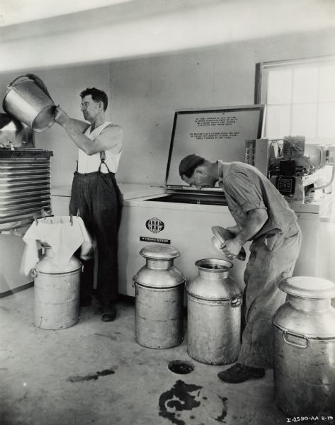 Two men are working with milk cans and buckets near a McCormick-Deering milk cooler. The original caption reads: "James McFarland," which may refer to the owner of the cooler shown.