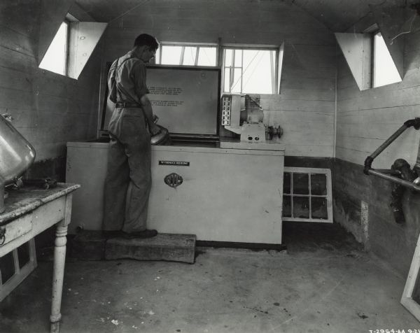 A man stands on a wooden step holding a milk can in a cooler.  Windows are on each wall. Original caption states: "McCormick-Deering 8-can milk cooler owned by Mrs. Manel Yates."