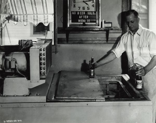 A man holds two bottles of beer on top of a cooler. A clock is on the wall with a sign that reads: "No Beer Sold After Midnite." Original caption states: "McCormick-Deering 6 can cooler owned by Otis Brandis Tavern."