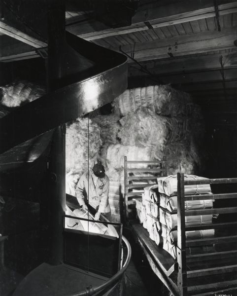 A factory worker loads bales of twine onto a platform, possibly at the McCormick Twine Mill. Original caption reads:  "The workman is shown here placing bales of twine into the shute[sic] which will carry it to the loading dock for shipment to all parts of the world."