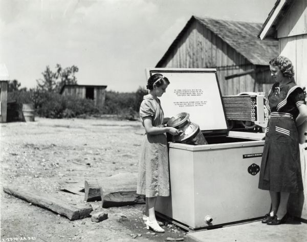 Two women standing by a milk cooler. One of the women is pulling out a milk can from the cooler and the other woman is leaning against a wall.  In the background are wood buildings.
