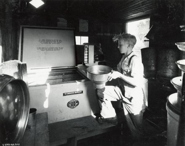 A boy in overalls stands with a foot on a step next to a milk cooler in a small room, near an open doorway.  Original caption states: "McCormick-Deering 6 can milk cooler owned by Mrs. H.A. Rechamp."
