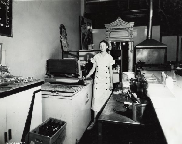 A woman is standing by a cooler with a beer bottle in her hand at Reed's Cafe near a stove and countertop. Original caption states: "McCormick-Deering 6 can milk cooler owned by D.J. Reed."