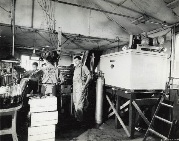 Three workers stand near a conveyor carrying bottles at the Pittsfield Bottling Works. A milk cooler sits on a platform nearby. The back of one worker's shirt reads "Hinie's Soda." Original caption states: "McCormick-Deering milk cooler installation owned by Pittsfield Bottling Works, W.H. Williamson."