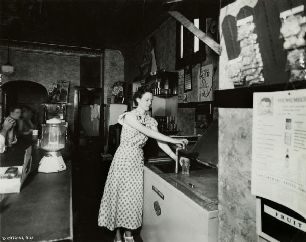A woman is filling a glass from a faucet mounted on a McCormick-Deering 6 can cooler in the Floyd Kaylor Restaurant. Three people are sitting at a counter on the left.