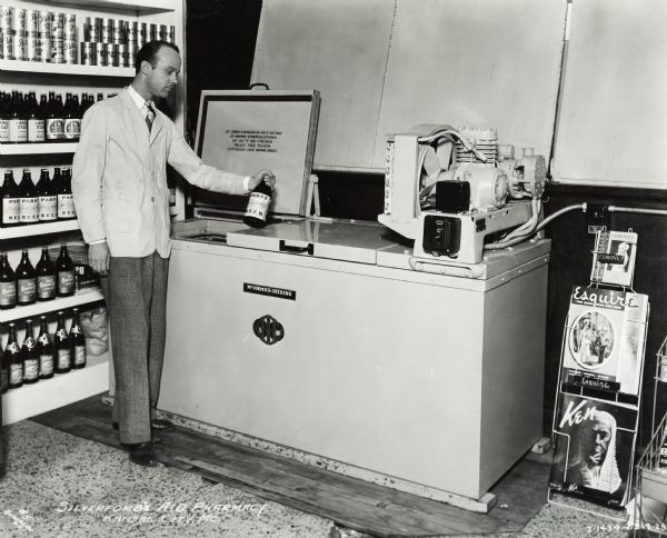 A man stands by a cooler with a bottle of Pabst beer in his hand. Bottles and cans of beer are along the wall behind him. On the right is a magazine stand with the magazines <i>Coronet</i>, <i>Esquire</i>, and <i>Ken</i>. Original caption states: "McCormick-Deering 4 in line beer cooler."