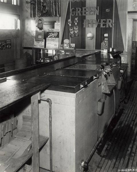 A McCormick-Deering ten can cooler sits underneath a counter next to the soda faucets at Clyde Cockershaw's Bar. In the background are advertisements for alcohol and cigarettes.