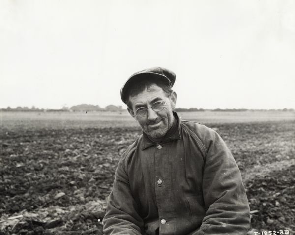 Fred Prante in a field wearing a hat, eyeglasses, and a coat. Original caption states: "Owner of Mc-D 4 can milk cooler."