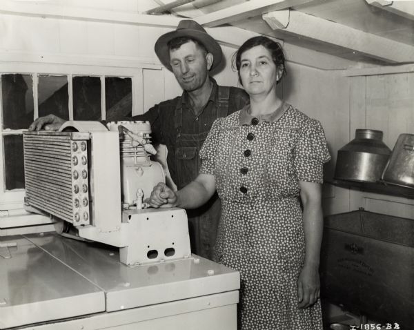 Mr. and Mrs. Edgar Schildknecht stand by a McCormick-Deering six can "engine drive" cooler in a small room.