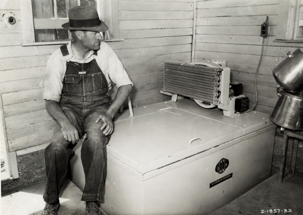 Charles A. Woodson, dressed in overalls, is sitting on the corner of a 6 can electric milk cooler inside a wooden building. Two metal pails are on the right.