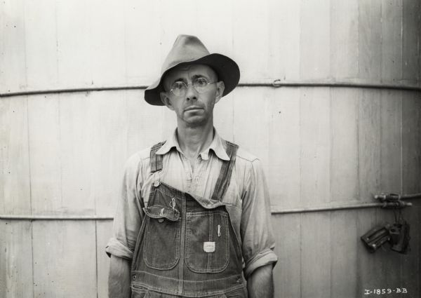 Farmer William A. Henke, dressed in hat, eyeglasses and overalls, and standing in front of a silo(?). Mr. Henke was the owner of a McCormick-Deering six can milk cooler.