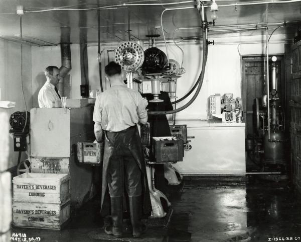 Workers standing at a machine putting bottles into crates. One of the workers is wearing an apron and rubber knee-high boots. A McCormick-Deering eight can milk cooler is in the right background. The crates are labeled "Davern's Beverages" and "Cobourg". The plant may have been located in Cobourg, Ontario, Canada.