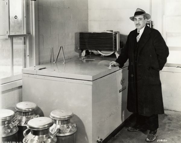 A man in a hat and long coat with rubber protectors over his shoes stands next to a McCormick-Deering cooler. Four milk cans are in the foreground.  Original caption states: "McCormick-Deering 8-can Milk Cooler owned by Dr. Benjamin J. Birk."