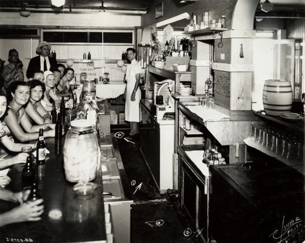 View down counter of women and men sitting and drinking beer or soda. Two men stand on the back left. One woman is wearing a bathing cap, and other women appear to be wearing bathing suits.  A man in an apron is working behind the counter. Original caption states: "4 Can-In-Line McCormick-Deering Cooler."