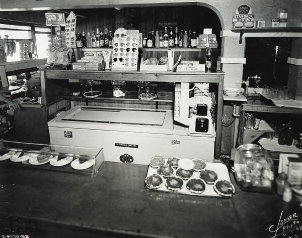 A customer's view of the lunch counter at the Cup & Saucer Restaurant. Pies and donuts(?) are on the counter. A 4 can in-line McCormick-Deering cooler is under displays of chewing gum, sunglasses and other items for sale.