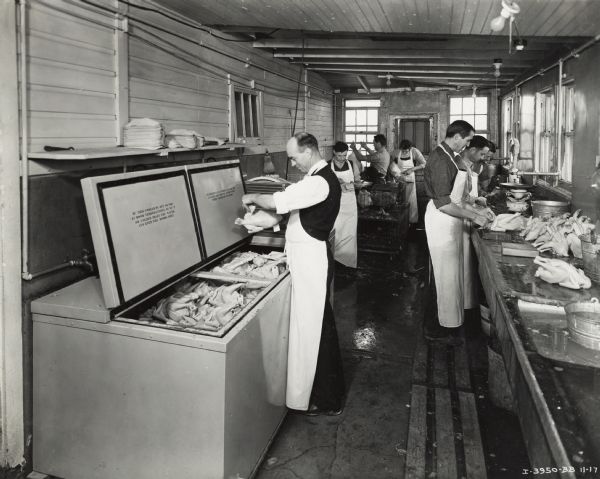 Workers wearing aprons are plucking, cleaning, and dressing chickens at the Schauls Live Poultry Farm. One man is looking at a chicken over an open McCormick-Deering cooler. Live chickens are in a cage in the middle. Original caption gives location as "6800 No. California Avenue.  Chicago Cooler Installation."