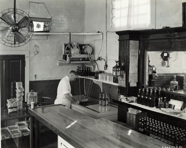 Elevated view pf W.T. Hening, dressed in an apron, leaning over a McCormick-Deering No. 4 beverage cooler at the Seventh Street Buffet. A bar or lunch counter is in the foreground, with bottles of beer on the mirrored bar in the background. There is a display case with cigars, potato chips and other items on the left. A large fan hangs from the ceiling. The original caption identifies the address of the establishment as "814 W. Seventh, Joplin, Mo."
