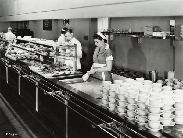 Workers stand behind a food service counter at the Tractor Works (factory) cafeteria. The counter is loaded with dishes, cups and saucers. One woman is moving a milk jar in a beverage cooler.