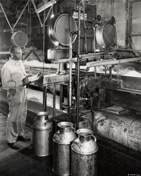 A factory worker stands with a clipboard and inspects a machine, possibly at the McCormick Twine Mill.  Caption on photograph reads: "This inspector is shown making tests to insure that the proper amounts of emulsion, oil, and repellents are being applied to the fiber."