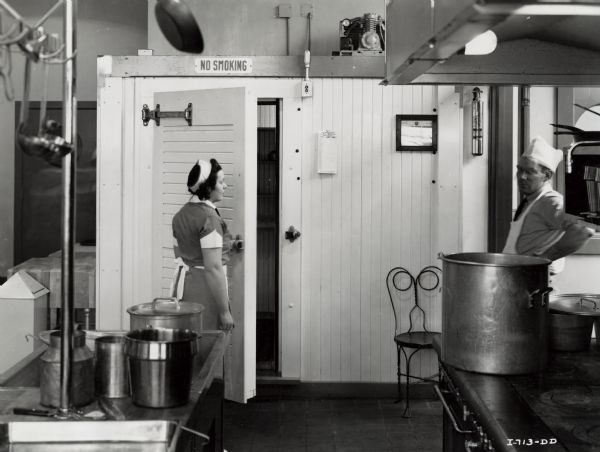 A woman in uniform standing by the open door of a walk-in-cooler in the cafeteria at International Harvester's Tractor Works (factory).  A man is standing on the right behind a stove. A "No Smoking" sign is above the door of the cooler.