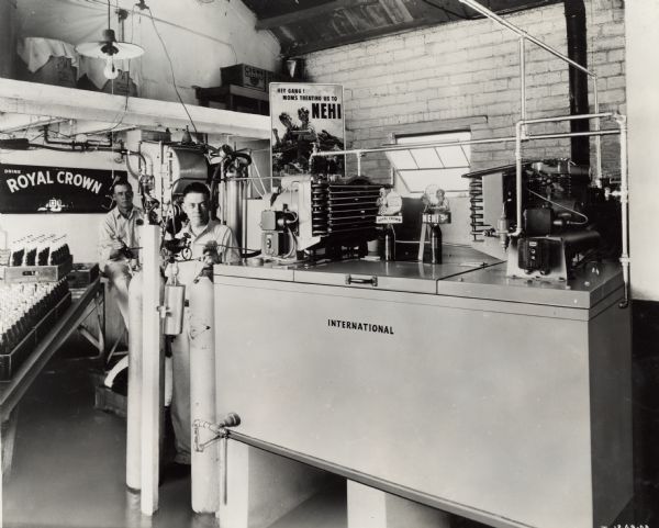 Workers standing near bottling equipment at the Royal Crown Cola Company. Advertising signs for Royal Crown and Nehi soda are in the background.