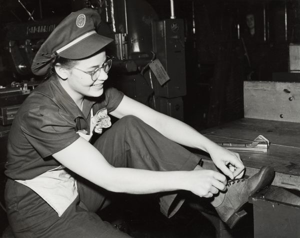 Factory worker Joan Hebrink with safety shoes. Original caption states: "Closed-toe safety shoes are required wear by all women employees of the plant. All employees also wear safety goggles for the protection of their eyes. In this photograph Joan Hebrink puts on her safety shoes prior to starting work." The factory may be the St. Paul Works.