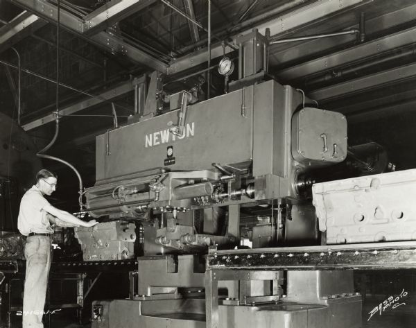 Factory worker near a conveyor belt at International Harvester's Indianapolis Works. The man has tattoos on both arms. Original caption states: "Newton Rise and Fall-Type machine for milling both sides of crankshaft bearings and lock slot in cylinder block. Note the conveyor on the same plane with the fixture on this machine. The clamping action is controlled by air, which makes the operation safer and easier."