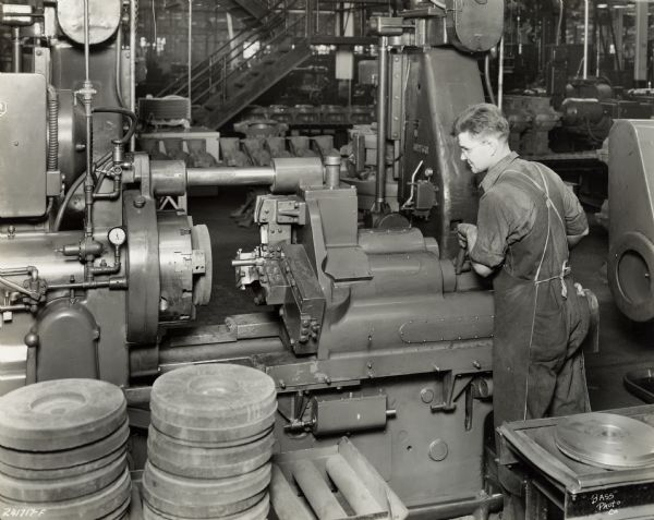 Factory worker in overalls and an apron at International Harvester's Indianapolis Works. Original caption states: "Radial-type Gisholt automatic machine for rough turning, facing, and boring the flywheel.  This machine is equipped with an air-operated chuck."