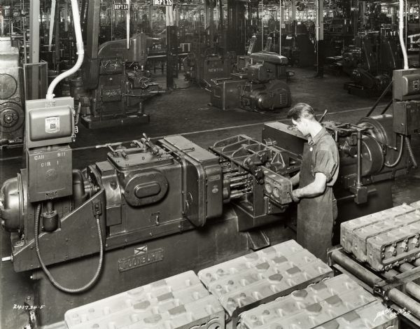 Factory worker operates a machine at International Harvester's Indianapolis Works. Near the top are two signs: "Dept. 15" and "Dept. 14". Original caption states: "Foote-Burt two-way machine for drilling all holes in top and bottom of cylinder heads as well as all cylinder head stud holes through the cylinder head. This method assures proper sizes and spacing of holes."
