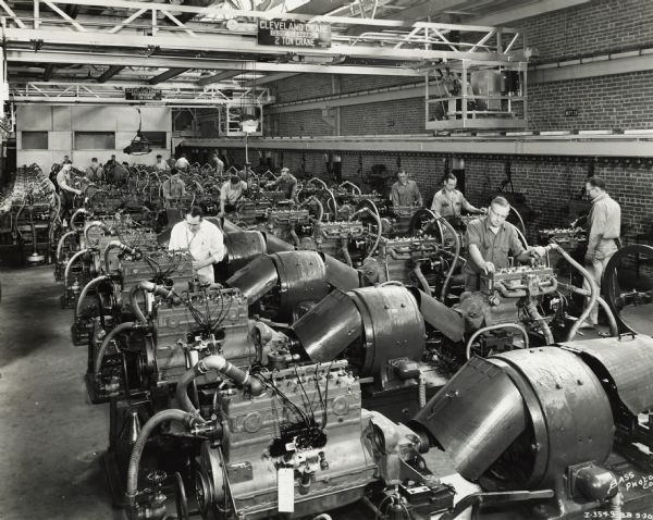 Factory workers tending to engines at International Harvester's Indianapolis Works. Other men are operating overhead cranes labeled "Cleveland Crane-Close Clearance-2 Ton Crane."