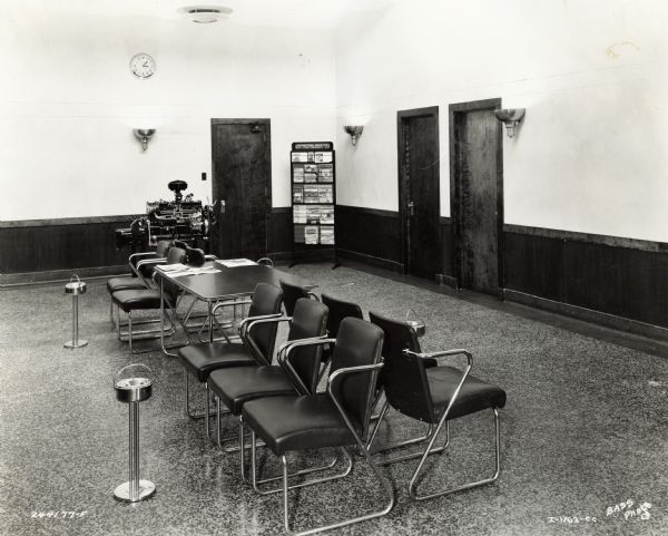 Ten chairs, a table, ashtrays, and a pamphlet and magazine display in the lobby of International Harvester's Indianapolis Works. An engine is on display in the back of the room. Original caption states: "Lobby in new Administration building. Furniture is chrome with blue leather upholstery."