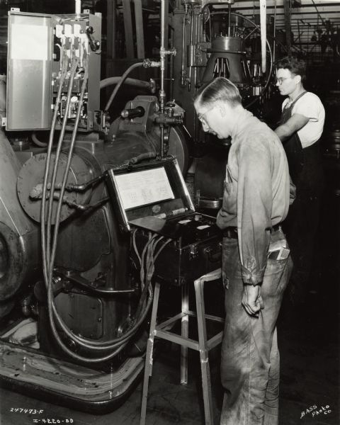 Factory workers at International Harvester's Indianapolis Works (Truck Engine Works). Original caption states: "Checking 10 H.P. motor on brake drum lathe with industrial analyzer. Note starter connections for analyzer, fuses are removed and fuses with leads permanently attached are inserted. This makes it possible to run a test with minimum loss of production time. Note safety guard over analyzer terminals."