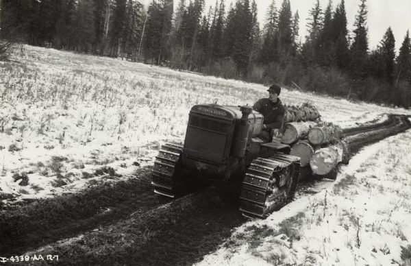 A man pulls logs with an International T-20 TracTracTor (crawler tractor) along a dirt road in a snow-covered field.