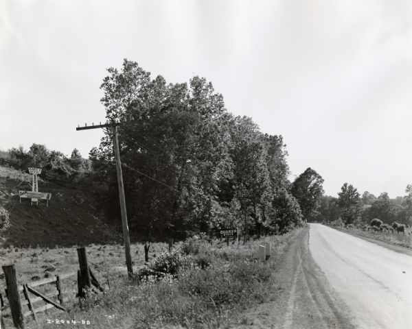 A country road including a barbed wire fence, advertising signs and trees. One of the signs is in the shape of an airplane diving toward the ground and advertises York & Massey, an International Harvester dealership. The sign reads: "Make a Dive for York and Massey.  Buick, Good Year, International Trucks and Tractors, Hardware." Another sign advertises: "Quaker State" oil.