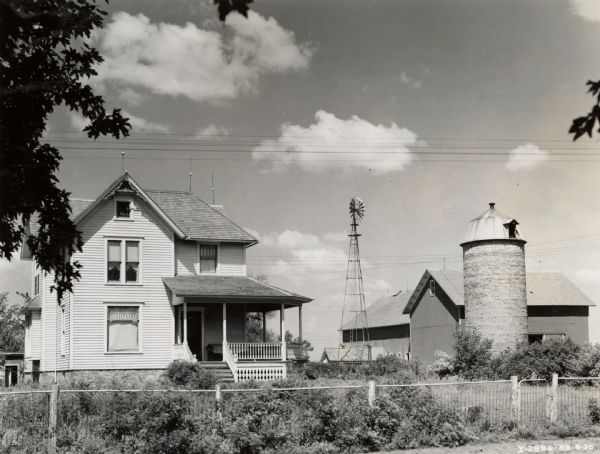 Front view of a farmhouse with a barn, silo, and windmill in the background.