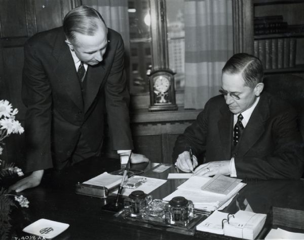 International Harvester President Sidney G. McAllister sits at a desk in his office while Comptroller C.E. Jarchow stands on the left.